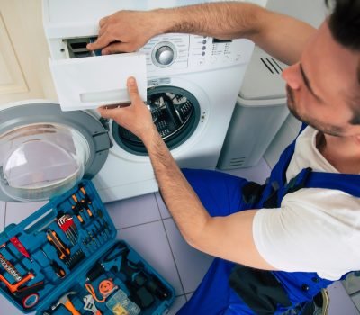 The young handsome repairman in worker suit with the professional tools box is fixing the washing machine in the bathroom